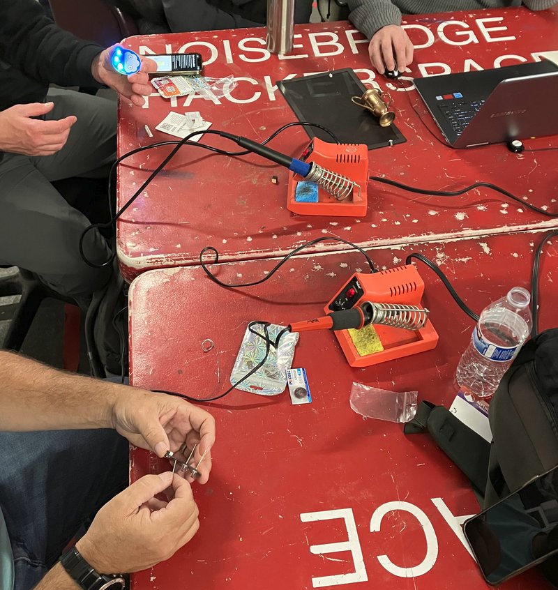 A shot of a red table with the lettering "NOISBRIDGE HACKERSPACE" There are soldering irons and electronic parts visible; people's hands are shown assembling electronic kits